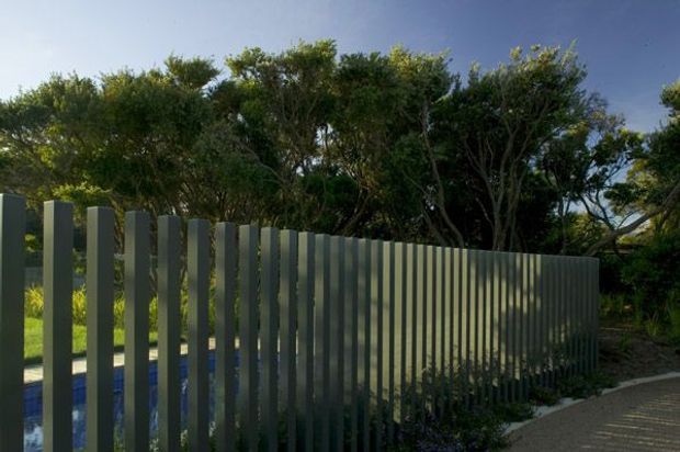 Exterior view of a contemporary vertical-slat privacy fence in a landscaped setting. The evenly spaced dark grey metal or painted posts screen a partially visible blue pool and garden, with mature trees behind and a paved walkway along the fence.