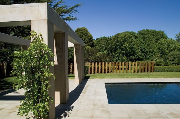 A landscaped outdoor pool area with a rectangular swimming pool edged in pale stone paving. A substantial concrete pergola or colonnade frames the left side, with climbing greenery, timber fencing, clipped hedges, mature trees, and a clear blue sky in the background. The scene has a clean, geometric contemporary garden design.