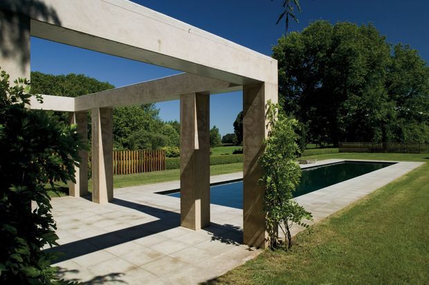 A landscaped outdoor pool area featuring a minimalist concrete pergola with strong rectangular beams and columns, pale tiled paving, and a long narrow swimming pool bordered by lawn and mature trees.