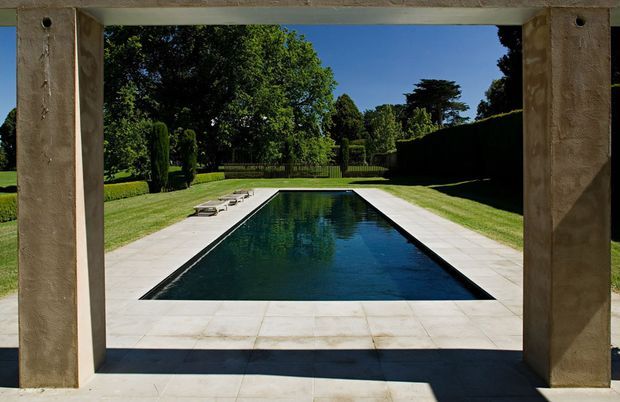 A formal rectangular swimming pool viewed through a substantial stone or concrete-framed opening. Pale paved coping surrounds the pool, with manicured lawns, clipped hedges, mature trees, and a dark metal gate creating a symmetrical garden setting. Strong sunlight and deep structural shadows give the scene a refined, classic outdoor character.