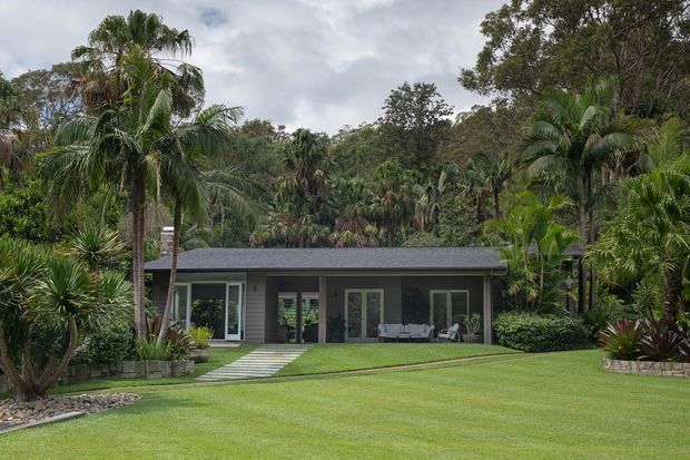 Single-storey contemporary residence set within a lush tropical landscape, featuring a low-pitched dark roof, dark horizontal exterior cladding, extensive glazed doors and windows, and a broad manicured lawn with a stepping-stone path leading to the house.