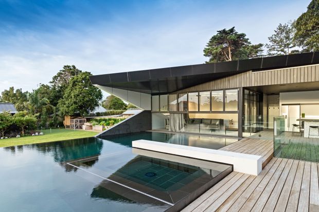 A contemporary single-storey residence with a dramatic cantilevered flat roof and extensive floor-to-ceiling glazing, opening onto a timber deck and large reflecting-style swimming pool. The home features dark exterior panels, pale vertical wall cladding, an outdoor built-in spa or shallow tiled pool, and a clean modern indoor-outdoor connection surrounded by landscaped greenery.