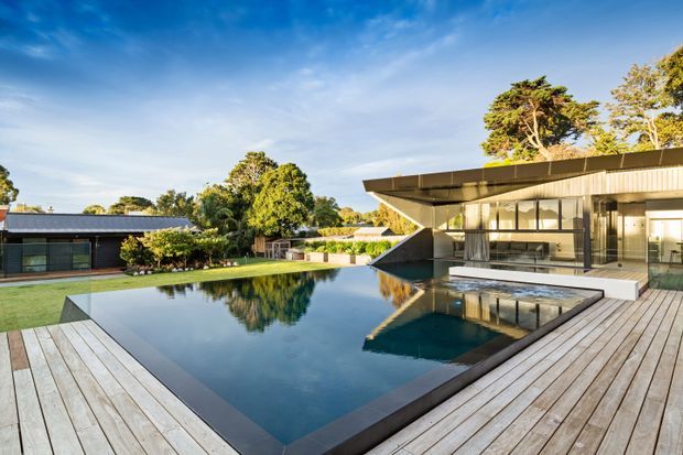A contemporary single-storey residence arranged around a large reflective swimming pool. The image shows broad timber decking, expansive glazing, a dramatic cantilevered flat roof, dark metal detailing, and landscaped grounds under a clear sky.