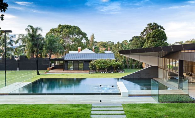 Contemporary residential property viewed from the landscaped backyard, featuring a long swimming pool, glass balustrades, a manicured lawn and stepping-stone path, a black-and-timber pavilion-like house with a broad low-pitched roof, and a separate dark-roofed structure with extensive glazing. A fenced tennis court and mature tropical planting are also visible.