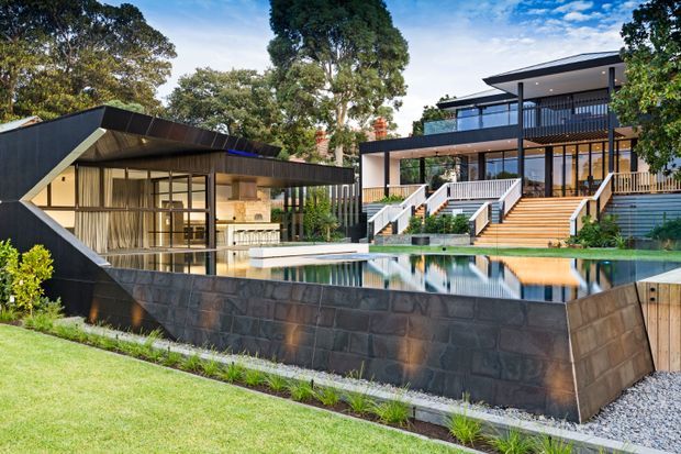 A contemporary multi-level residential property viewed from the landscaped grounds, with dark geometric cladding, extensive glazing, broad roof overhangs, terraces and balconies. A long reflective swimming pool occupies the foreground, with timber steps and decks connecting to the main house and a pavilion-like structure.