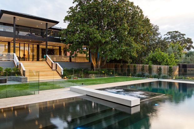 Contemporary multi-level residence with broad roof overhangs, extensive glazing, warm timber stair and deck elements, and dark horizontal cladding. The landscaped setting includes a large reflective swimming pool with a projecting white platform, glass pool fencing, manicured lawn, mature trees, and planted borders.