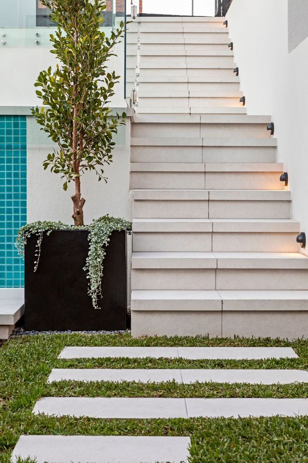 A contemporary outdoor courtyard stairway with broad light-coloured tiled or stone steps, integrated step lighting, and a glass balustrade. A black rectangular planter with a slender tree and trailing greenery sits beside the stairs, with turquoise tiled wall cladding visible to the left. Stepping slabs cross a neatly maintained grass lawn in the foreground.