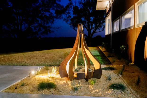 A dusk exterior view of a landscaped residential entry area, featuring a sculptural timber or rust-toned metal garden installation surrounded by gravel, grasses, and planted beds. A contemporary timber-clad house with illuminated windows and a covered walkway is visible on the right, with a lawn and dark tree line beyond.