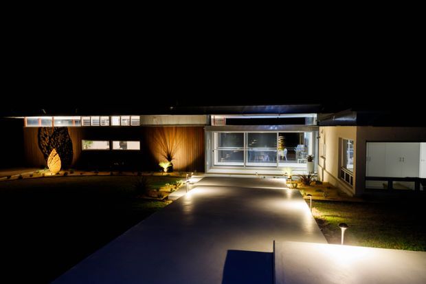 Night-time exterior view of a low, modern residential building approached by a broad illuminated concrete driveway or paved forecourt. The facade features extensive glazing, a long horizontal roofline, warm timber-toned wall panels, and landscaped garden beds with architectural lighting.