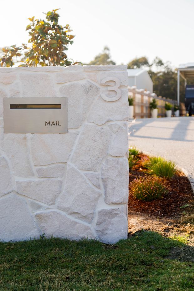 A close-up exterior view of a pale, irregularly textured stone-clad mailbox pillar marked with the number 3 and a brushed metal mail slot. A landscaped residential driveway, low planting, lawn, and part of a modern property are visible in the background.