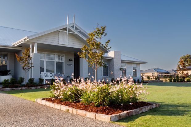 Exterior view of a single-storey suburban residence with a light weatherboard facade, gabled roof, prominent decorative gable detailing, white louvred shutters, and a covered front porch. The home is set behind a broad manicured lawn with a raised garden bed and gravel driveway.