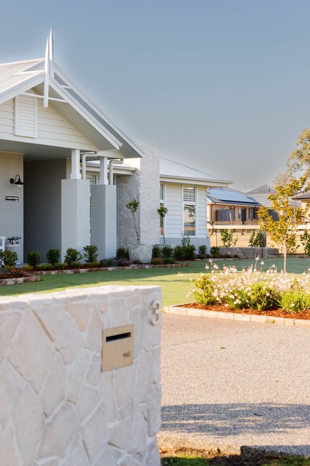 A bright residential streetscape showing a light-coloured single-storey home with a prominent gabled porch, white weatherboard cladding, decorative gable detailing, a pale stone feature wall, landscaped front garden, lawn, driveway, and a stone mailbox pillar in the foreground.