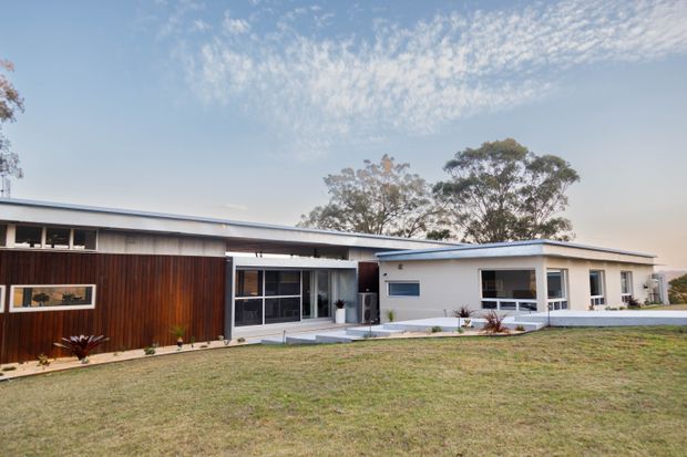 Single-storey modern residential house viewed from the front across a broad lawn. The building has a low-pitched roof, long horizontal proportions, white rendered walls, dark timber vertical cladding, extensive dark-framed glazing, and a recessed central entry or courtyard connection. Mature trees are visible behind the home under a bright blue sky.