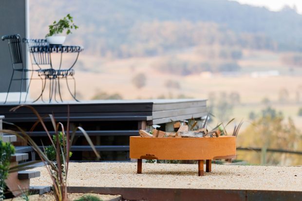 A contemporary outdoor terrace or deck overlooking a broad rural, hilly landscape. The foreground features a gravel-finished terrace, dark horizontal decking or bench structures, delicate metal outdoor furniture with a potted plant, and a rust-toned rectangular fire pit or planter. The scene has a relaxed, minimalist setting with muted natural colours and expansive views.