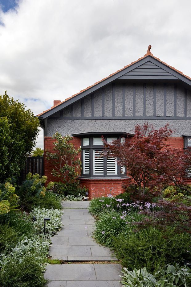 Front exterior view of a traditional gabled brick house, with grey rendered and half-timbered upper facade, decorative gable detailing, dark-framed windows with white shutters, and a landscaped garden path leading to the entrance. The planting is dense and ornamental, creating a welcoming residential streetscape.
