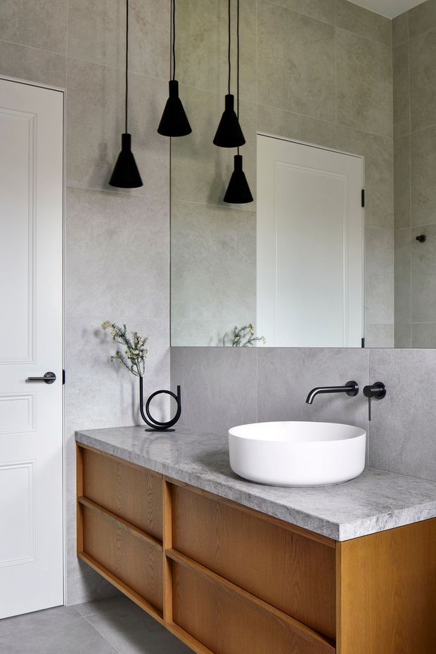 Contemporary bathroom vanity featuring a floating warm timber cabinet with large drawers, a grey stone countertop, white vessel basin, matte black wall-mounted tapware, grey tiled walls and floor, a large mirror, and four black pendant lights. White paneled doors are visible at either side.