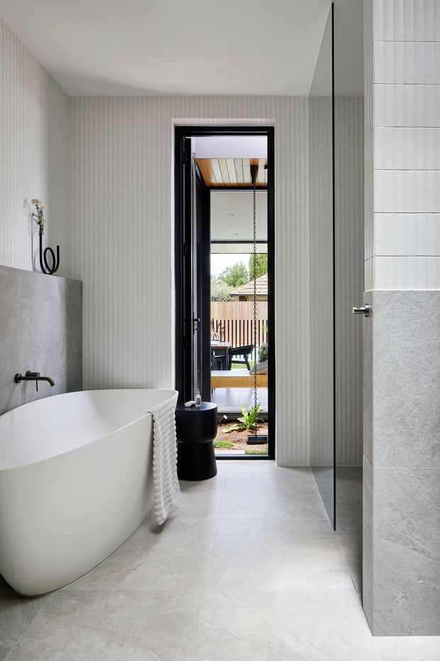 A bright, contemporary bathroom featuring a freestanding white bathtub, wall-mounted black tapware, pale vertically fluted wall tiles, and large-format light grey stone-look floor and wall finishes. A frameless glass shower screen occupies the right side, while a black-framed glazed door opens to an outdoor deck or courtyard. The room has a clean, refined, spa-like atmosphere with strong natural light.