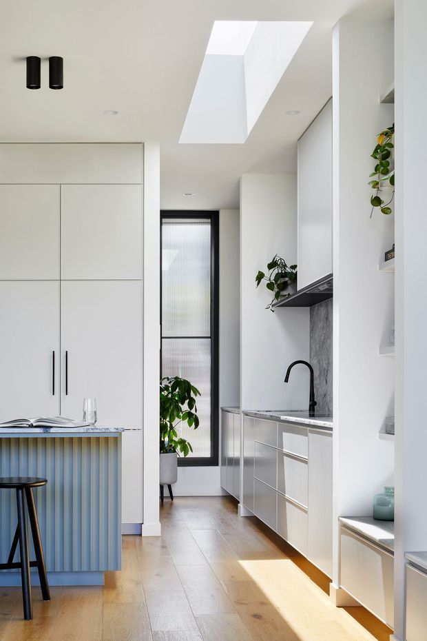 A bright, narrow contemporary kitchen with white full-height cabinetry, integrated appliances, a grey stone backsplash and benchtop, and a black gooseneck tap. A blue fluted island with a dark stool occupies the foreground, while timber flooring, a tall translucent window, indoor plants, and a large skylight add warmth and natural light.