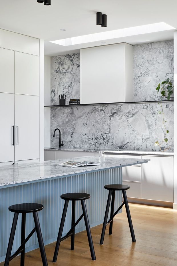 A bright contemporary kitchen with extensive white cabinetry, a dramatic grey-veined stone splashback and benchtops, a large island with vertical fluted detailing, and three black bar stools. A skylight and black cylindrical pendant fixtures add contrast, while timber flooring and a trailing indoor plant provide warmth.