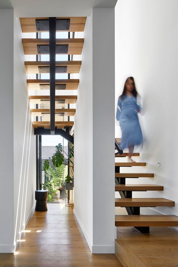 A bright, narrow modern stair hall with floating timber stair treads and matching overhead ceiling slats, supported by a dark central steel spine. White walls and structural columns frame the space, with warm timber flooring and a glazed opening at the end containing indoor plants. A blurred person is ascending the stairs.