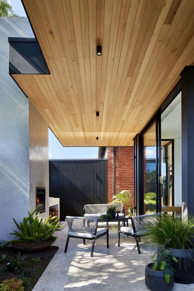 A refined covered outdoor courtyard patio adjoining a contemporary home. The space features a warm timber-lined soffit with recessed downlights, large black-framed glazed doors, a red brick wall, dark vertical metal fencing, a built-in outdoor fireplace, concrete paving, and modern wire-frame lounge furniture surrounded by potted greenery.