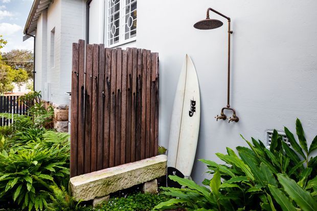 A coastal-style exterior side yard beside a white-painted masonry building, featuring an outdoor copper-toned shower, a weathered vertical timber privacy screen, a rough stone bench, and a surfboard. Lush green planting surrounds the area, with a barred window and pitched roof edge visible.