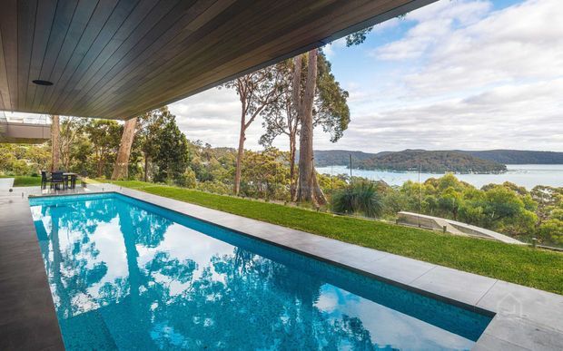 A modern covered outdoor pool terrace overlooking a calm coastal or lakeside waterway. The image features a long rectangular swimming pool, dark timber soffit, pale concrete pool surround, manicured lawn, native landscaping, and distant wooded hills across the water.