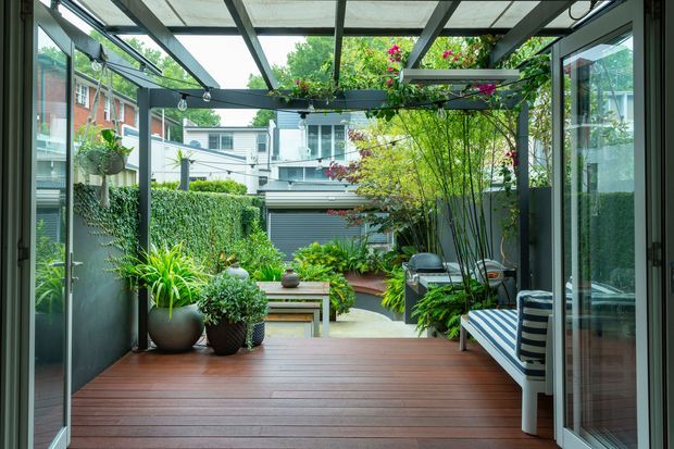 A covered outdoor deck or courtyard terrace with warm timber decking, dark metal framing, extensive glass doors and a translucent roof. The space is furnished with a striped built-in bench and surrounded by lush potted and planted greenery, opening toward a landscaped urban residential courtyard and garage.
