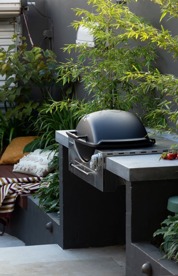 A compact landscaped outdoor entertaining area with a built-in stainless-steel barbecue grill, dark cabinetry, stone-paved flooring, raised planter boxes, bamboo and leafy greenery, and a cushioned bench nook.