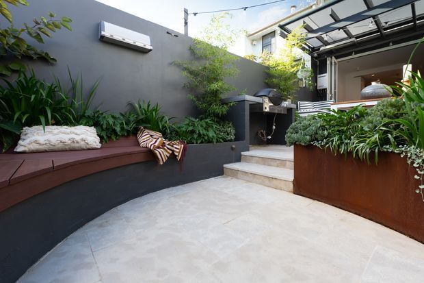 A contemporary landscaped courtyard or patio with pale stone paving, dark rendered walls, built-in timber bench seating, raised planters, lush foliage, and steps leading to a covered outdoor entertaining area. A barbecue or outdoor kitchen is visible near the rear, with a glass-and-metal awning over the adjoining opening.