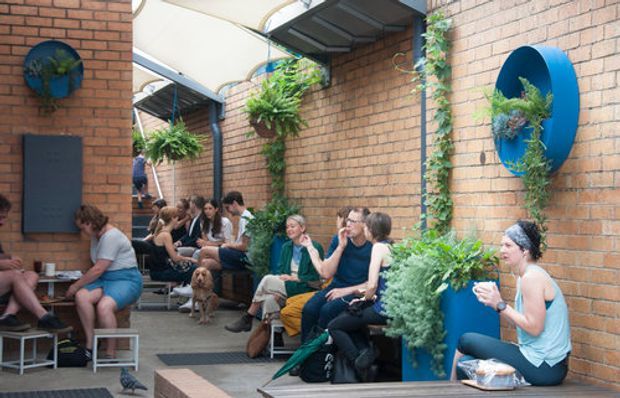 A lively semi-enclosed brick courtyard or outdoor seating area with exposed brick walls, concrete paving, overhead translucent canopy panels, integrated benches, and numerous hanging and wall-mounted plants. People are seated and socialising along the perimeter, with strong blue circular planter features adding colour.