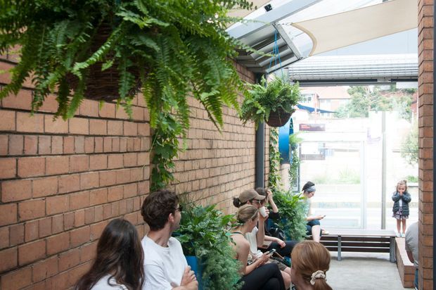 A covered, semi-open brick-lined courtyard or walkway with built-in bench seating, hanging ferns, climbing plants, and several people gathered along the seating area. A translucent canopy and glazed opening provide shelter and daylight, creating a green, communal outdoor space.