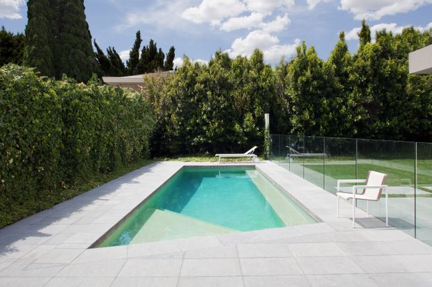 A modern rectangular swimming pool set within a private landscaped courtyard or garden. The pool is surrounded by pale tiled paving, dense hedging, transparent glass fencing, and simple white outdoor lounge chairs. The scene is bright and clean with a contemporary outdoor design.