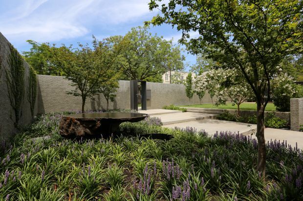 A landscaped contemporary courtyard garden with textured rendered masonry walls, mature trees, clipped hedges, ornamental grasses and flowering groundcover. A shallow dark stone water feature or sculptural basin sits among the planting, with pale paved paths and steps leading through the space.