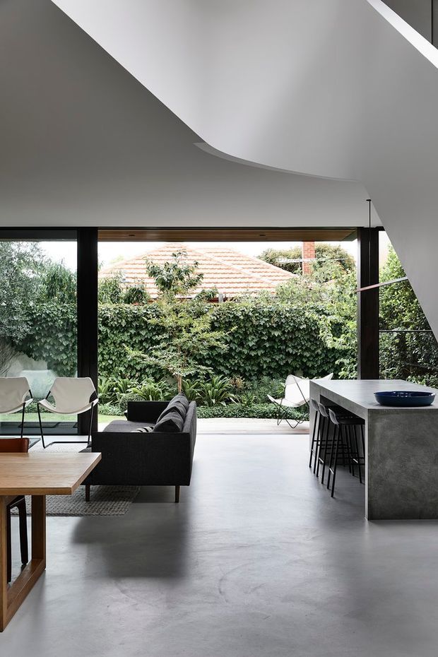 A refined open-plan living and kitchen space with a sculptural curved white ceiling, full-height black-framed glazing, polished grey concrete flooring, and a minimalist concrete island. The room opens onto a lush enclosed garden, with a dark sofa, low timber coffee table, white lounge chairs, and black bar stools creating a calm contemporary interior.