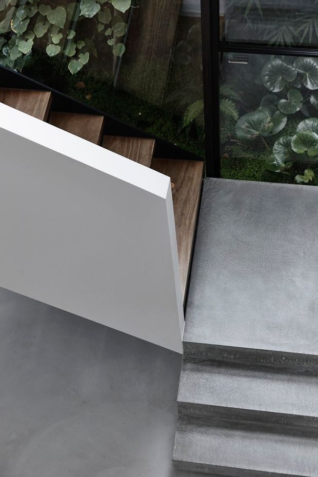 A close-up architectural detail of a contemporary stairway, featuring dark timber stair treads, a crisp white geometric balustrade or wall, and grey concrete flooring and steps. Black-framed glazing beside the stairs reveals dense planted greenery, creating a strong indoor–outdoor connection. The composition is minimalist and materially focused.