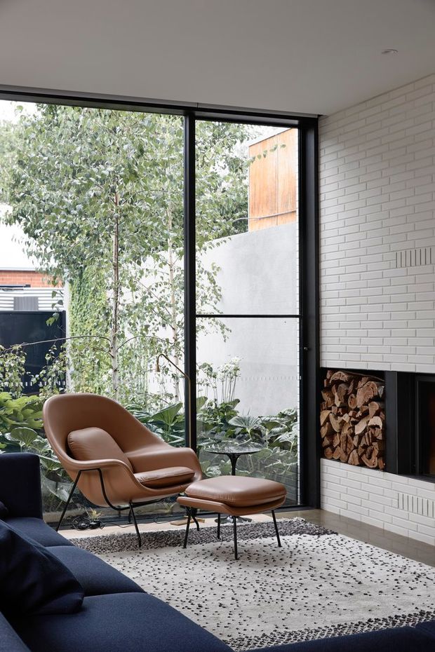 A contemporary living room with a floor-to-ceiling black-framed glazed wall overlooking a planted courtyard. The room features a white painted brick fireplace wall with an integrated timber log niche, a deep navy sectional sofa, and a tan leather lounge chair with matching ottoman. A textured pale rug and indoor greenery create a refined, natural atmosphere.