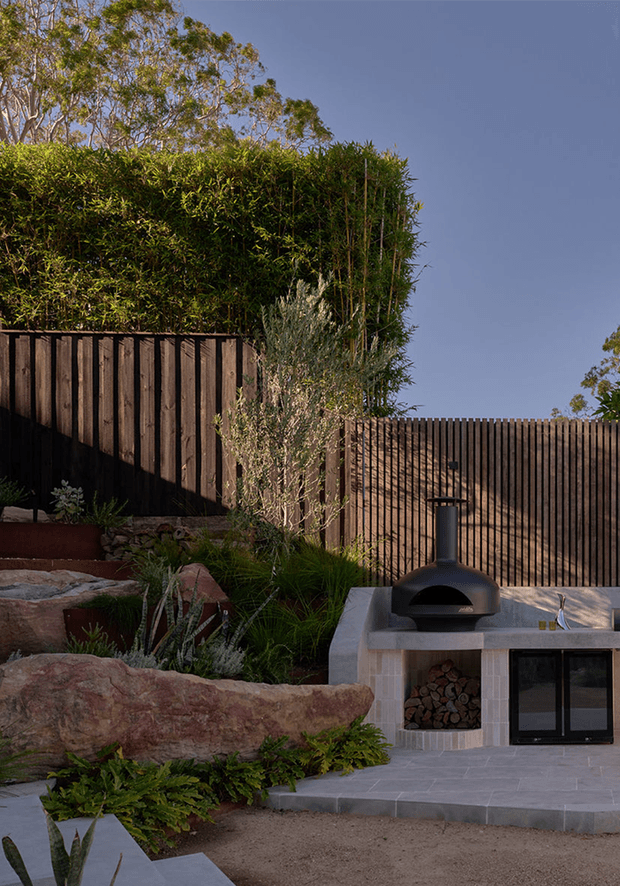 A landscaped outdoor entertaining area with a built-in masonry outdoor kitchen. The space features a dark timber slat privacy fence, dense hedging, large natural boulders, layered drought-tolerant planting, pale paved flooring, a concrete benchtop, a black dome pizza oven, and integrated firewood storage.