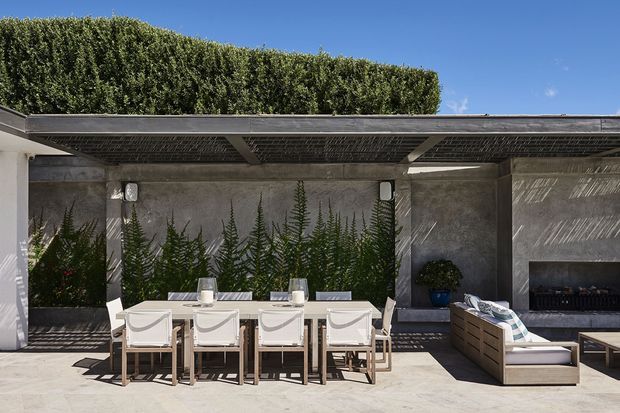 A sophisticated outdoor entertaining area beneath a broad pergola, featuring a long timber dining table with upholstered chairs, a built-in garden wall with tall greenery, and a cushioned outdoor lounge beside a dark built-in fireplace or feature wall. Concrete surfaces and a slatted overhead structure create strong patterned shadows in bright sunlight.