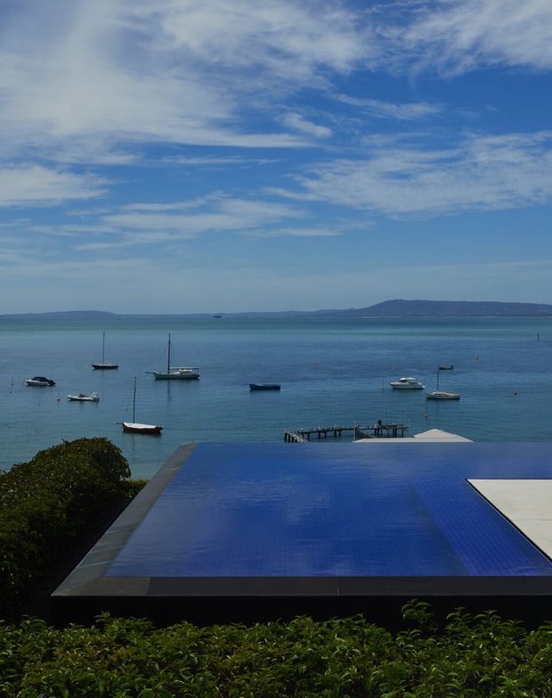 A serene coastal view from an elevated property, dominated by a dark-edged rectangular infinity pool extending toward calm blue water. Several moored boats, a small pier, distant low hills, and a bright partly cloudy sky create a tranquil waterfront setting. Dense green planting borders the foreground pool terrace.