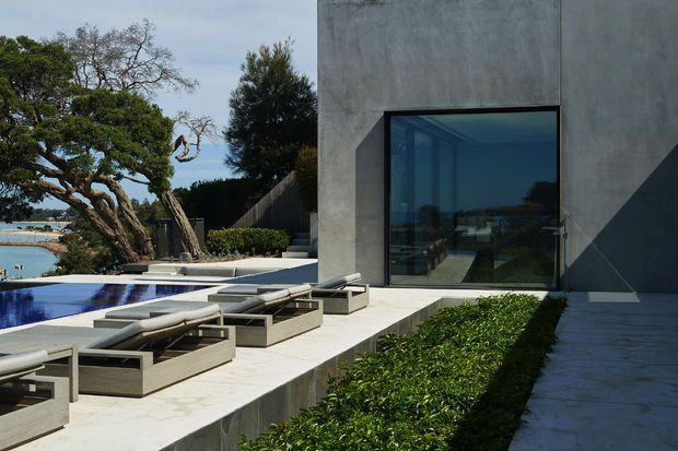 Contemporary concrete-clad residence opening onto a pool terrace, with a large dark-framed glazed opening, pale stone paving, built-in sun loungers, clipped hedges, and mature coastal landscaping. Water and shoreline are visible beyond the garden.