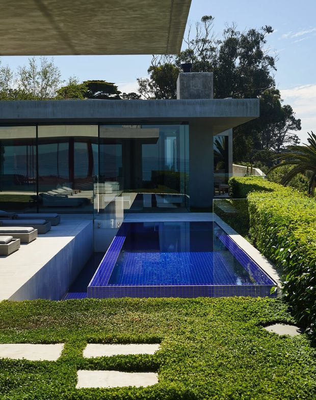 A modern concrete residence with extensive floor-to-ceiling glazing, a flat projecting roof, and a rectangular swimming pool clad in vivid blue mosaic tiles. Pale outdoor loungers sit beside the pool, while clipped hedges and layered landscaping frame the terrace and stepping-stone path. The image has strong geometric lines, reflective glass, and bright natural daylight.