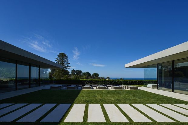 A modern low-rise residential property arranged around a landscaped central lawn, with two minimalist flat-roofed wings, extensive floor-to-ceiling glazing, dark structural frames, and broad pale paving slabs forming a geometric path. Outdoor lounge seating is visible beyond the lawn, with a distant coastal horizon.