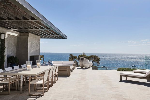 A spacious elevated outdoor terrace overlooking the ocean, with a long dining table and chairs beneath a broad slatted roof overhang, a built-in or freestanding outdoor kitchen area, lounge seating, and pale stone or concrete paving. The scene has a clean, contemporary coastal character with expansive sea views.
