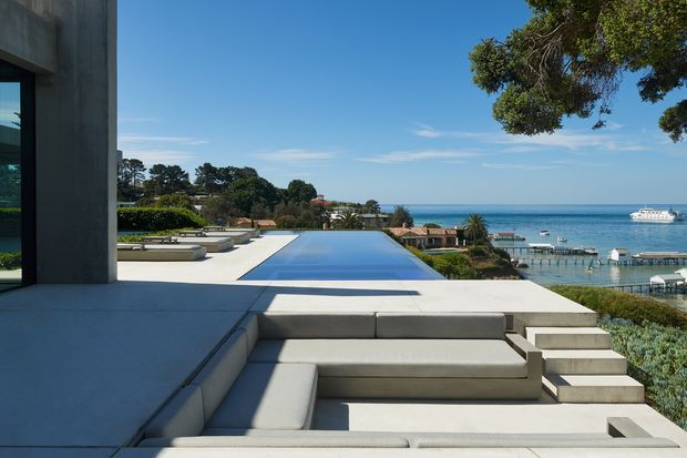 Contemporary coastal residence viewed from an elevated outdoor terrace, featuring a long infinity-edge pool, built-in concrete seating, broad pale paving, and stepped levels overlooking the ocean, marina, and anchored boats. The composition emphasizes clean horizontal lines, substantial concrete forms, and a bright blue-sky waterfront setting.