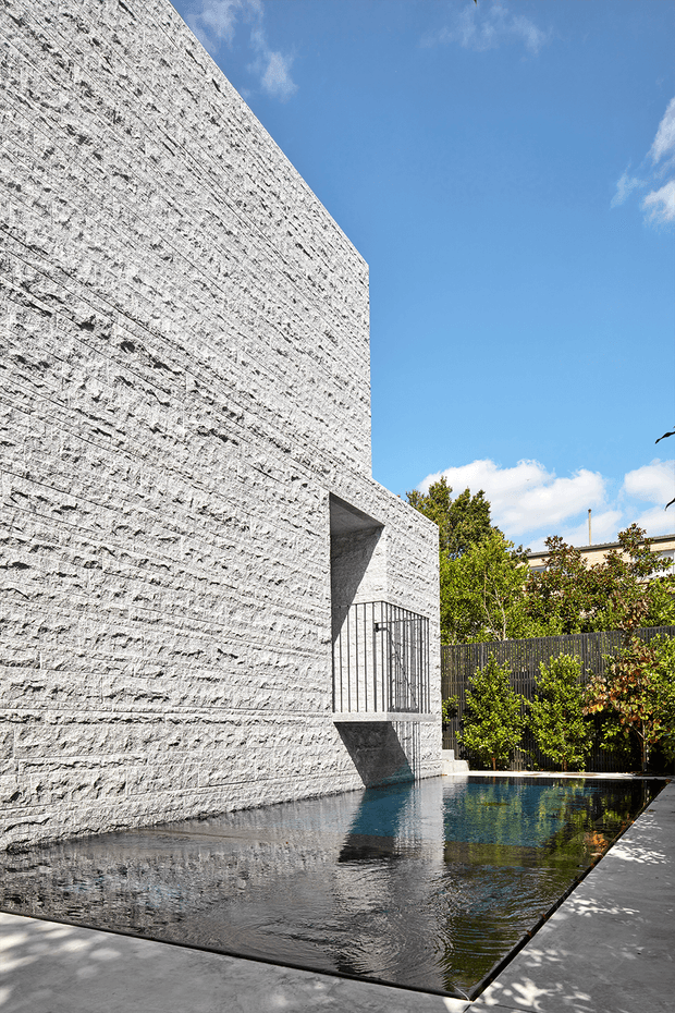 A stark contemporary exterior featuring a tall white textured masonry wall with a recessed, gated opening overlooking a narrow reflective pool. Dark fencing, greenery, and a paved edge surround the water, creating a minimalist courtyard-like setting beneath a bright blue sky.