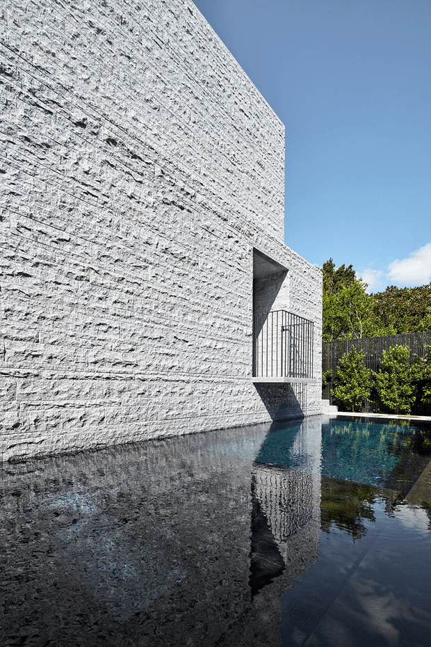 A minimalist modern exterior featuring a tall, monolithic white textured masonry wall beside a dark reflective swimming pool. A recessed balcony or entry opening with black metal railing interrupts the wall, while dense green planting and fencing frame the pool edge under a clear blue sky.