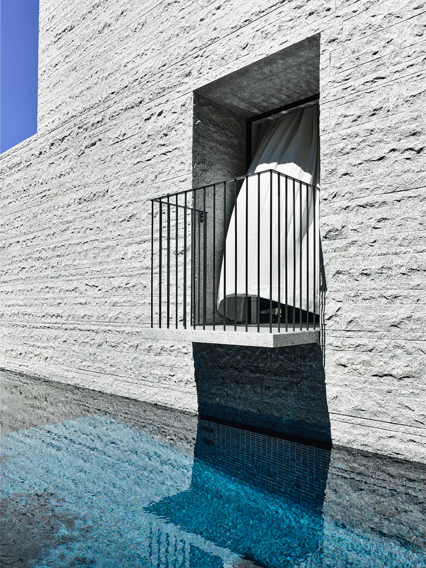 A stark architectural exterior detail showing a small projecting balcony with a slim black metal balustrade, recessed doorway, and draped curtain. The facade is clad in heavily textured pale stone or concrete, with strong sunlight casting deep shadows. A vivid blue-tiled reflecting pool or water feature occupies the foreground, creating a high-contrast, minimalist composition.