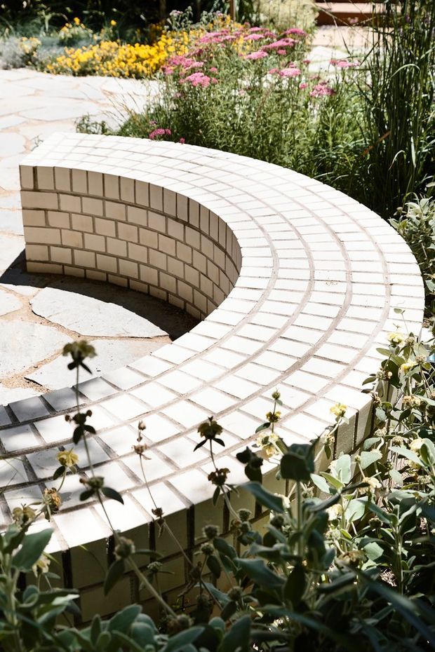A landscaped outdoor garden with a curved, semi-circular low masonry bench or retaining wall finished in small pale rectangular tiles. The curved surface surrounds a recessed paved area, with irregular stone paving and abundant flowering and ornamental planting around it. The scene is sunlit and organic, with a refined garden-design character.