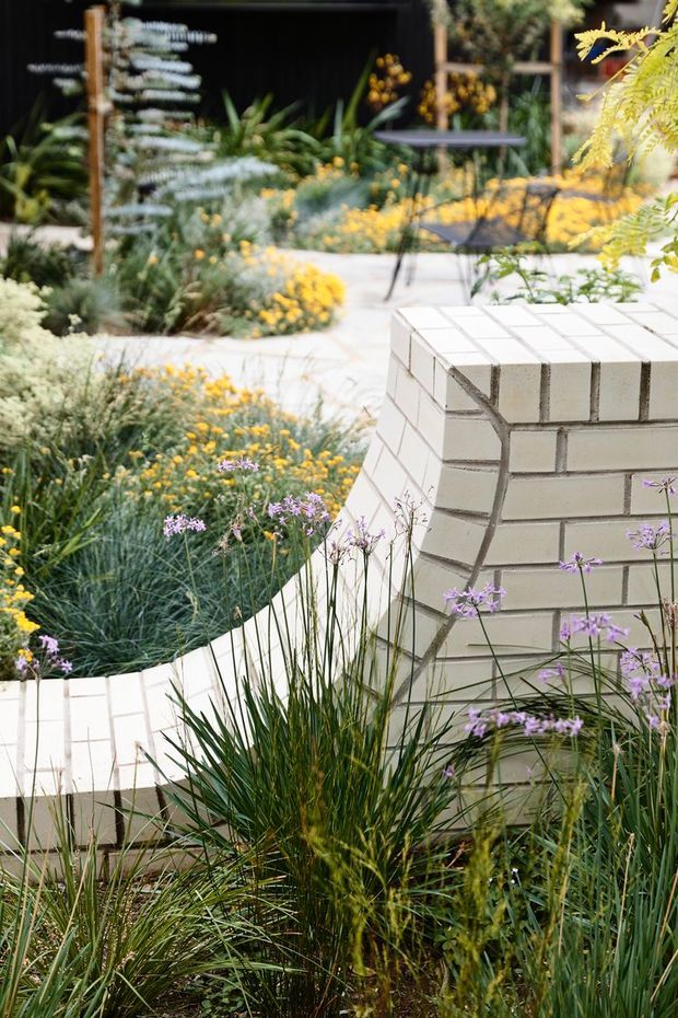 A landscaped outdoor courtyard or garden with curved pale paving and a low white brick masonry structure, surrounded by ornamental grasses, yellow flowering plants and purple blooms. A small dark outdoor table and chairs sit in the background near a dark-clad garden wall or building.